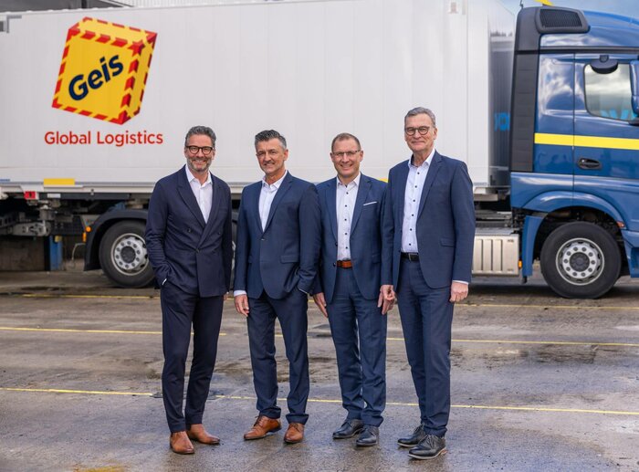 Vier Männer in Business-Anzügen vor einem Lkw mit Geis-Logo
